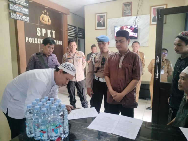 Seorang pria mengenakan kopiah putih sedang menyerahkan dokumen di kantor polisi dengan beberapa orang di sekitarnya.