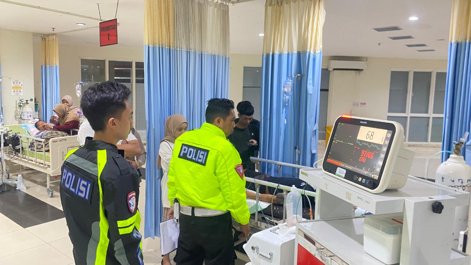 Dua petugas polisi berdiri di dekat peralatan medis di rumah sakit, dengan pasien di tempat tidur di latar belakang. Dua petugas polisi berdiri di dekat peralatan medis di rumah sakit, dengan pasien di tempat tidur di latar belakang.