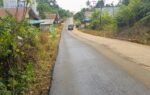 Jalan aspal yang menghubungkan area pedesaan dengan kendaraan melintas di tengah pepohonan.