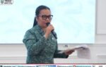 Wanita berokupasi batik warna teal berkacamata sedang membawakan mic sambil memegang dokumen, latar belakang layar putih, logo Pemerintah Kabupaten Murung Raya terlihat di pojok kiri atas, foto diambil oleh Rid