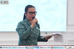 Wanita berokupasi batik warna teal berkacamata sedang membawakan mic sambil memegang dokumen, latar belakang layar putih, logo Pemerintah Kabupaten Murung Raya terlihat di pojok kiri atas, foto diambil oleh Rid