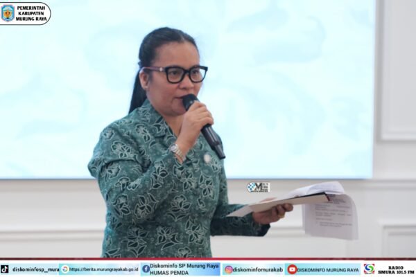 Wanita berokupasi batik warna teal berkacamata sedang membawakan mic sambil memegang dokumen, latar belakang layar putih, logo Pemerintah Kabupaten Murung Raya terlihat di pojok kiri atas, foto diambil oleh Rid
