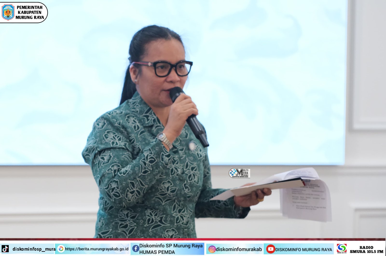 Wanita berokupasi batik warna teal berkacamata sedang membawakan mic sambil memegang dokumen, latar belakang layar putih, logo Pemerintah Kabupaten Murung Raya terlihat di pojok kiri atas, foto diambil oleh Rid
