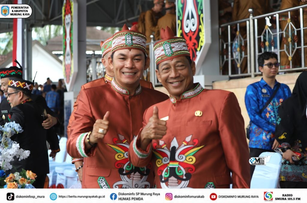 Heriyus dan Rahmanto Muhidin tersenyum sambil mengacungkan jempol, mengenakan busana adat merah saat menghadiri HUT ke-76 Barito Utara.