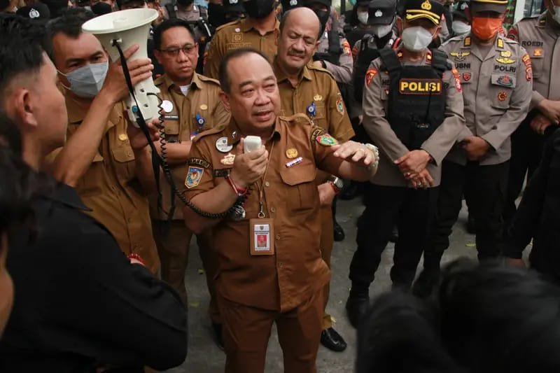 Seorang pejabat berbaju dinas cokelat berbicara sambil memegang perangkat komunikasi di tengah warga dan petugas berseragam.