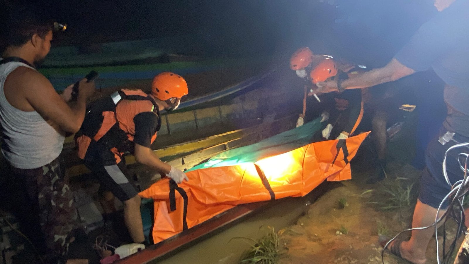 Petugas SAR berhelm oranye mengevakuasi Bakir dengan tandu oranye di tepi sungai pada malam hari setelah ditemukan tewas tenggelam.