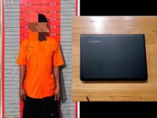 Seorang pria berbaju oranye berdiri di depan papan ukur, di samping laptop Lenovo hitam yang tergeletak di atas meja kayu.