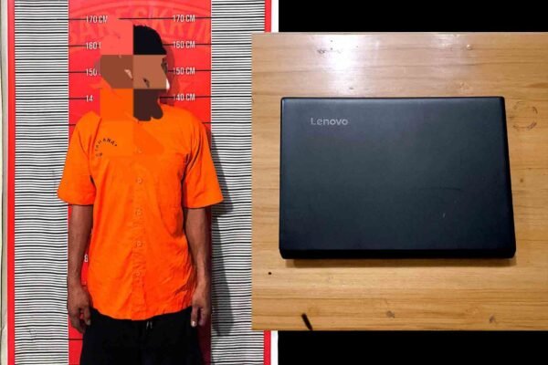 Seorang pria berbaju oranye berdiri di depan papan ukur, di samping laptop Lenovo hitam yang tergeletak di atas meja kayu.