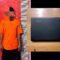 Seorang pria berbaju oranye berdiri di depan papan ukur, di samping laptop Lenovo hitam yang tergeletak di atas meja kayu.